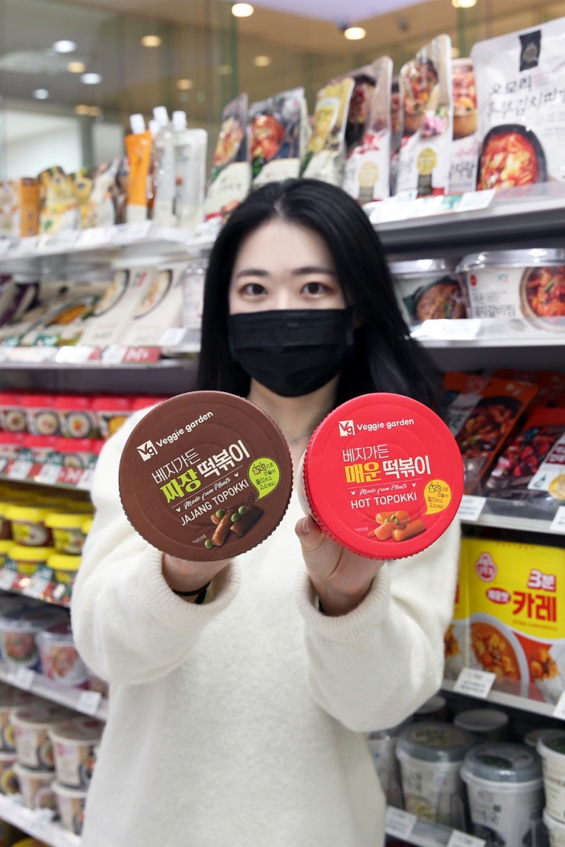 (사진=GS리테일) GS25 매장의 간편식 매대 앞에서 모델이 비건간편식 짜장떡볶이와 매운떡볶이를 들고 있다