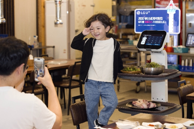 LG유플러스는 고객과 함께 ‘2030 부산 세계박람회(부산 엑스포)’ 유치를 응원하는 다양한 이벤트를 전개했다고 23일 밝혔다. 사진은 새벽숯불가든 부산대신점 고객이 부산 엑스포 유치 응원 판넬이 부착된 U+서빙로봇과 함께 기념사진을 촬영하는 모습. (사진 = LG유플러스 제공)