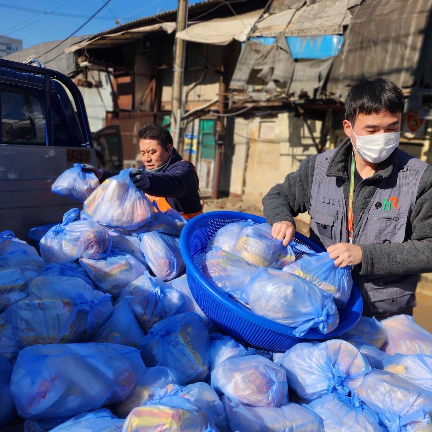 HR그룹 소속 봉사자들이 영등포 쪽방마을 이웃들에게 야외에서 따뜻한 떡국을 대접하고, 생필품이 담긴 선물을 정성스럽게 정리하여 전달할 준비를 하고 있다./HR그룹 미디어팀